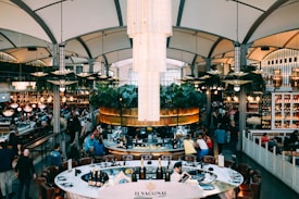 An upscale restaurant interior with a large elegant chandelier at the center, surrounded by lush greenery and people dining at various tables. The space features contemporary architecture with high ceilings and large windows. Shelves with bottles and decor line the walls, and the lighting creates a warm, inviting ambience.