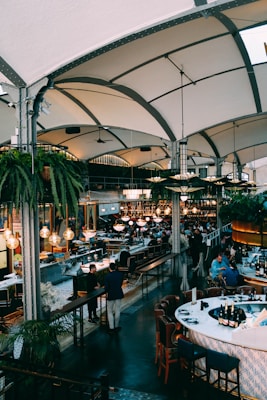 A large, bustling indoor restaurant with high arched ceilings and lush green hanging plants. The space is filled with people sitting at tables and a bar, engaged in conversation. The lighting is warm, provided by multiple round chandeliers. The decor includes a modern combination of wood and metal with comfortable seating.