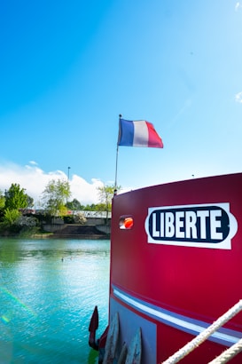 A vibrant blue sky and a calm body of water are seen with a boat at the forefront, which features a sign with the word 'Liberte'. A French flag is prominently displayed atop the boat. Trees and a wall in the background add a touch of greenery, while the water gently reflects the sky's hue.