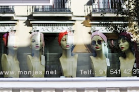 Mannequin heads are displayed in a shop window, each adorned with different hats and accessories, set against a backdrop reflecting buildings and trees from an urban street.