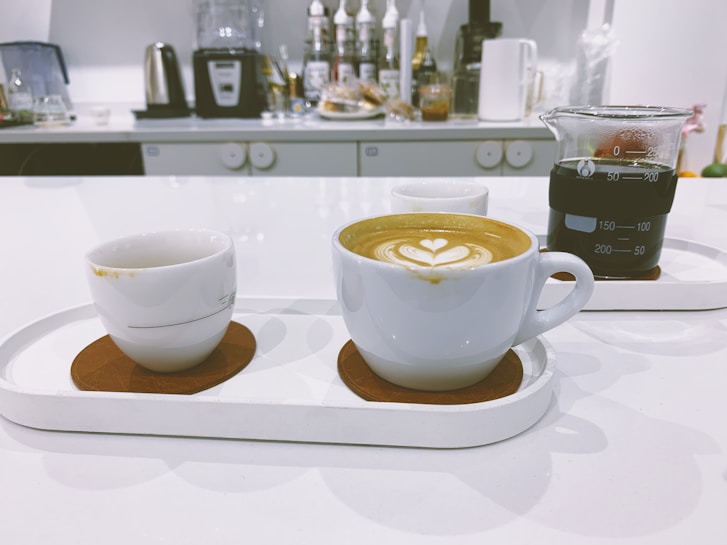 A modern kitchen counter with three cups arranged on trays. The central cup features a latte with heart-shaped latte art. To the right, a glass beaker filled with black coffee is visible. Various kitchen appliances and syrups are seen in the background.