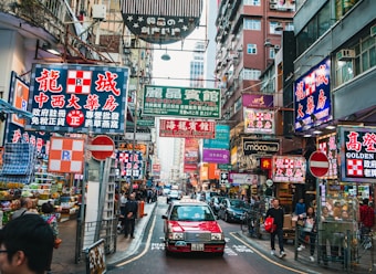 A bustling urban street scene filled with vibrant neon signs in both Chinese and English. Tall buildings line the narrow street, which is crowded with people walking along the sidewalks. A red taxi is driving down the road, and various storefronts and market stalls are visible, showcasing an energetic and lively city atmosphere.