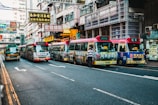 A bustling urban street lined with colorful minibuses displaying advertisements and traditional Chinese signage. The street is flanked by high-rise buildings with numerous signs, indicating a busy commercial district. Pedestrians are present, with one person standing near the minibuses, suggesting active public transportation.