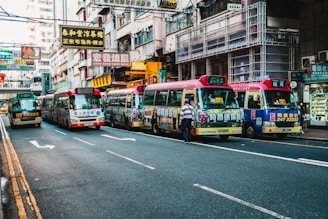 A bustling urban street lined with colorful minibuses displaying advertisements and traditional Chinese signage. The street is flanked by high-rise buildings with numerous signs, indicating a busy commercial district. Pedestrians are present, with one person standing near the minibuses, suggesting active public transportation.