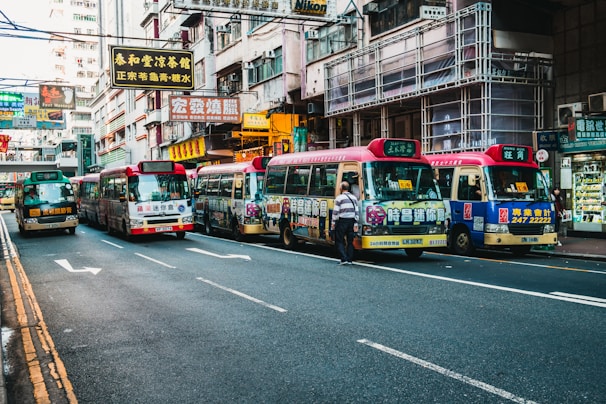 A bustling urban street lined with colorful minibuses displaying advertisements and traditional Chinese signage. The street is flanked by high-rise buildings with numerous signs, indicating a busy commercial district. Pedestrians are present, with one person standing near the minibuses, suggesting active public transportation.