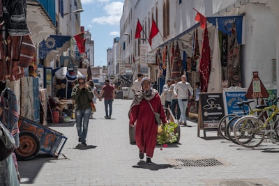 A bustling narrow street is lined with shops displaying textiles and goods. People walk along, with one person carrying a barrel-like object filled with greenery. Flags hang above, and bicycles are parked on the side. The atmosphere is lively and full of local culture.