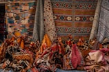 An assortment of colorful woven textile rugs, showcasing intricate patterns and various geometric designs. The rugs are displayed vertically and are hanging against a backdrop, suggesting a market or store setting.