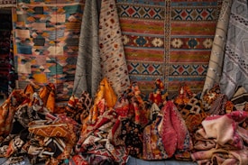 An assortment of colorful woven textile rugs, showcasing intricate patterns and various geometric designs. The rugs are displayed vertically and are hanging against a backdrop, suggesting a market or store setting.