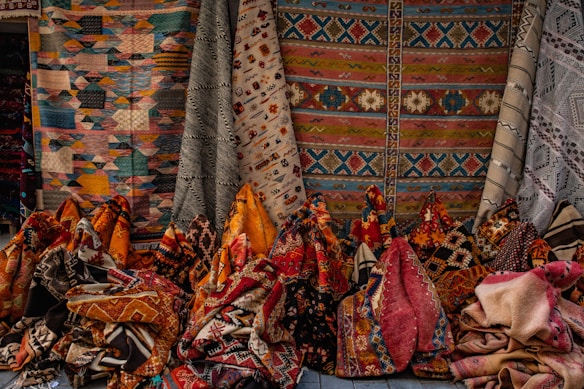 An assortment of colorful woven textile rugs, showcasing intricate patterns and various geometric designs. The rugs are displayed vertically and are hanging against a backdrop, suggesting a market or store setting.