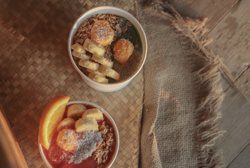 Two smoothie bowls are placed on a textured surface. The top bowl contains granola, banana slices, a dollop of nut butter, and coconut flakes. The bottom bowl features a bright red smoothie topped with banana slices, orange wedges, granola, and coconut flakes. A rustic burlap cloth adds a homely touch to the scene.