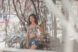 A young woman stands confidently in a garden-like setting, surrounded by twisting branches and foliage. She wears a floral crop top and distressed denim shorts, with her hand resting on her hip. The background features sunlight filtering through trees and some blurred elements in the foreground.