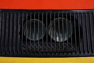 A close-up view of a metal ventilation grille with vertical slats, showcasing two circular openings behind the grille. The background features bright colors, including red and yellow.