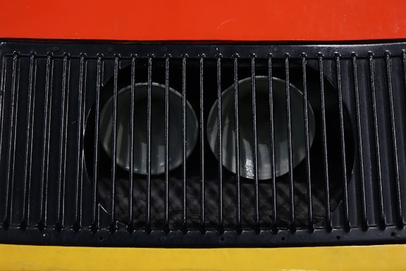 A close-up view of a metal ventilation grille with vertical slats, showcasing two circular openings behind the grille. The background features bright colors, including red and yellow.