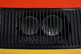 A close-up view of a metal ventilation grille with vertical slats, showcasing two circular openings behind the grille. The background features bright colors, including red and yellow.
