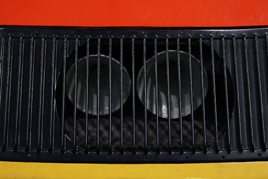 A close-up view of a metal ventilation grille with vertical slats, showcasing two circular openings behind the grille. The background features bright colors, including red and yellow.