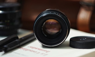 A camera lens is placed horizontally on a white book with red text. Nearby, there's a lens cap and a black pen. The background is blurred, focusing attention on the lens.