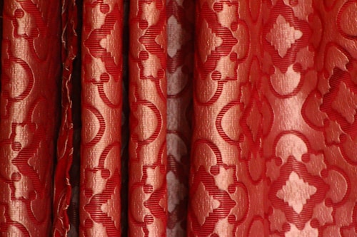 Detailed red fabric with an intricate geometric pattern. The design includes a series of repeating diamond and curved shapes, creating an elegant and luxurious appearance.