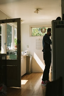A well-lit kitchen with wooden floors and a person standing near a refrigerator. Sunlight streams through a window, casting shadows on the hardwood floor. The kitchen counter is visible with a sink and various kitchen items. There is a window with a decorative glass design.