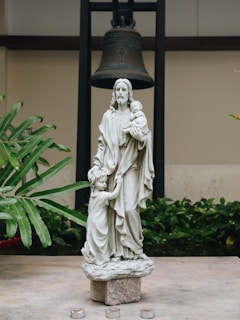 A stone statue depicting a figure holding a child in its arms and a young child at its side. The sculpture is set on a pedestal with a large bell hanging in the background. Lush green plants are visible to the side, and small candle holders are placed in front of the pedestal.