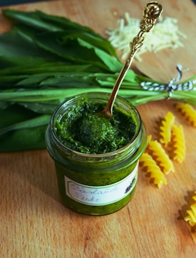 A jar of green pesto with a decorative spoon is placed on a wooden surface. Fresh green leaves are in the background, along with uncooked fusilli pasta and a garnish of grated cheese.