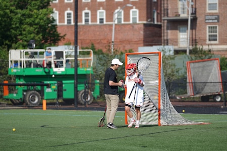 A lacrosse player wearing a red helmet and white uniform stands in front of a lacrosse goal, holding a lacrosse stick. Another person, wearing a black shirt and beige pants, is standing beside the player, possibly giving instructions or encouragement. In the background, a green utility vehicle is visible along with residential buildings and some trees.