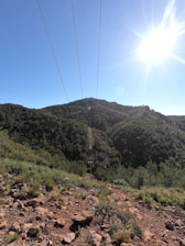 Power lines stretch across a scenic landscape featuring lush green hills and a clear blue sky. The sun shines brightly in the top right corner, casting light over the terrain. The foreground shows a rocky and grassy area, leading into a densely forested hill in the background.