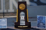 A large trophy stands prominently on a table, featuring the NCAA logo at the top and labeled '2019 Division I Lacrosse Championship' at the center. The base of the trophy reads 'National Champion.' On either side are informational signs about ticket sales for the championship event, with promotional images and details.