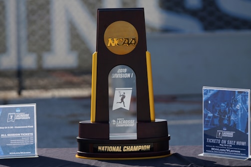 A large trophy stands prominently on a table, featuring the NCAA logo at the top and labeled '2019 Division I Lacrosse Championship' at the center. The base of the trophy reads 'National Champion.' On either side are informational signs about ticket sales for the championship event, with promotional images and details.