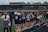 A group of female lacrosse players in navy uniforms stand on the sideline of a sports field. Some of them are holding lacrosse sticks and wearing helmets. Coaches and team staff are also present, and the background includes a modern sports facility with large windows. The players have their backs to the camera, and the scene conveys a sense of anticipation.