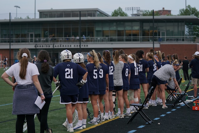 A group of female lacrosse players in navy uniforms stand on the sideline of a sports field. Some of them are holding lacrosse sticks and wearing helmets. Coaches and team staff are also present, and the background includes a modern sports facility with large windows. The players have their backs to the camera, and the scene conveys a sense of anticipation.