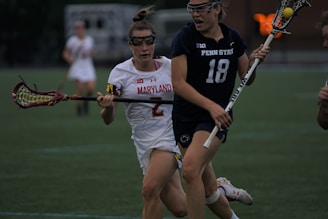Two female lacrosse players are in action on a sports field, with one wearing a white uniform marked 'Maryland' and the other in a dark uniform labeled 'Penn State'. Both are holding lacrosse sticks and appear to be competing for the ball.