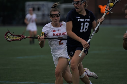 Two female lacrosse players are in action on a sports field, with one wearing a white uniform marked 'Maryland' and the other in a dark uniform labeled 'Penn State'. Both are holding lacrosse sticks and appear to be competing for the ball.