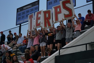 A group of young fans are sitting on stadium bleachers, holding up large signs with letters that spell 'TERPS'. They appear enthusiastic and are supporting a team. The background shows banners displaying years of football championships. Spectators around them vary in age, showing interest in the event.