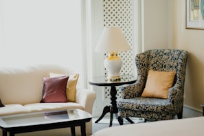 A cozy and elegant living space featuring a plush white sofa with decorative pillows in various colors, alongside a classic blue patterned armchair with a golden pillow. A round glass-topped side table with a white lamp rests between the seating. The setting includes soft natural light filtering in through a window with a decorative lattice.