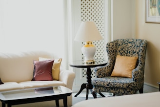 A cozy and elegant living space featuring a plush white sofa with decorative pillows in various colors, alongside a classic blue patterned armchair with a golden pillow. A round glass-topped side table with a white lamp rests between the seating. The setting includes soft natural light filtering in through a window with a decorative lattice.