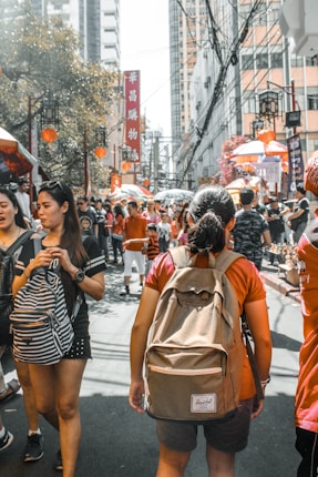 A bustling street scene featuring a large crowd of people walking among colorful market stalls and red lanterns. High-rise buildings line the background, with various advertisements and decorative elements enhancing the lively atmosphere.