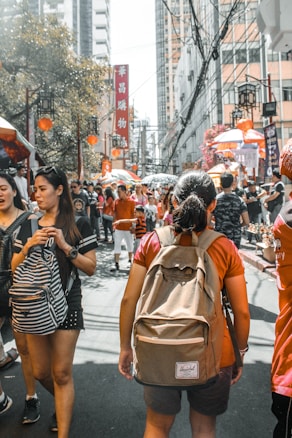 A bustling street scene featuring a large crowd of people walking among colorful market stalls and red lanterns. High-rise buildings line the background, with various advertisements and decorative elements enhancing the lively atmosphere.