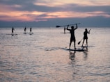 Several people are paddleboarding on a calm body of water during sunset. The silhouettes of the paddleboarders are visible against the backdrop of the colorful sky, which features shades of pink, purple, and blue. Some paddleboarders are holding their paddles above their heads while others are paddling.