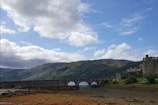 A historic castle is situated to the right, overlooking a broad landscape with a stone bridge spanning across a river or loch. The surrounding terrain includes rolling green hills under a partly cloudy blue sky.