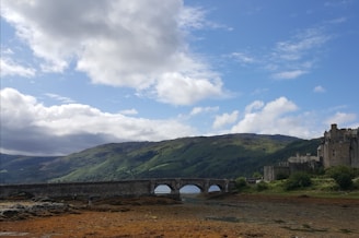 A historic castle is situated to the right, overlooking a broad landscape with a stone bridge spanning across a river or loch. The surrounding terrain includes rolling green hills under a partly cloudy blue sky.