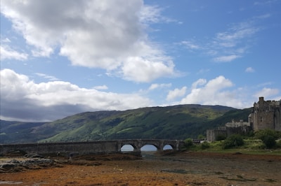 A historic castle is situated to the right, overlooking a broad landscape with a stone bridge spanning across a river or loch. The surrounding terrain includes rolling green hills under a partly cloudy blue sky.
