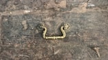 A rustic, weathered wooden surface features an ornate, vintage brass handle in the center. The wood is marked with scratches and discoloration, giving it an aged appearance. The handle has a floral motif with intricate detailing.