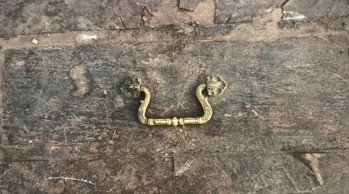 A rustic, weathered wooden surface features an ornate, vintage brass handle in the center. The wood is marked with scratches and discoloration, giving it an aged appearance. The handle has a floral motif with intricate detailing.