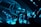 An indoor music event featuring a DJ booth illuminated with vibrant blue laser lights and large visual screens displaying abstract shapes. The crowd is silhouetted against the bright stage, adding to the energetic atmosphere.