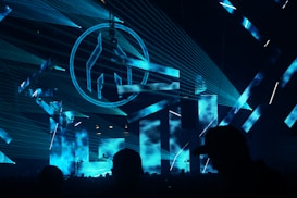 An indoor music event featuring a DJ booth illuminated with vibrant blue laser lights and large visual screens displaying abstract shapes. The crowd is silhouetted against the bright stage, adding to the energetic atmosphere.
