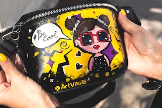 A hand-painted bag showing a cartoonish girl with sunglasses and a speech bubble saying '#Be Cool'. The background includes vibrant yellow, purple, and black colors, with decorative elements like stars and swirls.