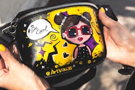 A hand-painted bag showing a cartoonish girl with sunglasses and a speech bubble saying '#Be Cool'. The background includes vibrant yellow, purple, and black colors, with decorative elements like stars and swirls.