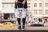 A person holding a tote bag with an artistic design featuring a girl with blue sunglasses and hair buns. The bag is white with colorful accents, including red and blue paint drips. The background shows a busy urban street scene with blurred pedestrians and a moving yellow car.