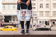 A person holding a tote bag with an artistic design featuring a girl with blue sunglasses and hair buns. The bag is white with colorful accents, including red and blue paint drips. The background shows a busy urban street scene with blurred pedestrians and a moving yellow car.