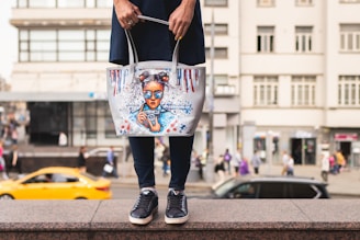 A person holding a tote bag with an artistic design featuring a girl with blue sunglasses and hair buns. The bag is white with colorful accents, including red and blue paint drips. The background shows a busy urban street scene with blurred pedestrians and a moving yellow car.
